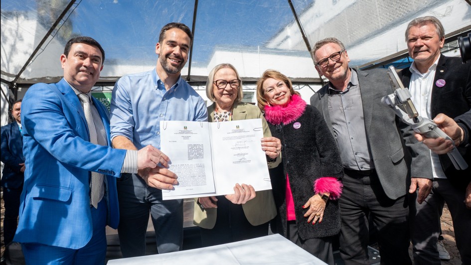 A fotografia mostra seis pessoas em uma celebração ao ar livre. O governador Eduardo Leite, de camisa azul de botão, e a secretária da Saúde, Arita Bergmann, de óculos e com um casaco bege claro, seguram um documento assinado. Eles sorriem. Do lado esquerdo, um homem de terno azul-claro com gravata estampada aponta para o documento. Do lado direito, uma mulher com um casaco preto, um cachecol rosa peludo e um homem de terno cinza sorriem. Um outro homem, à direita, também acompanha a cena. O evento ocorre sob uma tenda transparente, com luz natural.