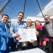 A fotografia mostra seis pessoas em uma celebração ao ar livre. O governador Eduardo Leite, de camisa azul de botão, e a secretária da Saúde, Arita Bergmann, de óculos e com um casaco bege claro, seguram um documento assinado. Eles sorriem. Do lado esquerdo, um homem de terno azul-claro com gravata estampada aponta para o documento. Do lado direito, uma mulher com um casaco preto, um cachecol rosa peludo e um homem de terno cinza sorriem. Um outro homem, à direita, também acompanha a cena. O evento ocorre sob uma tenda transparente, com luz natural.