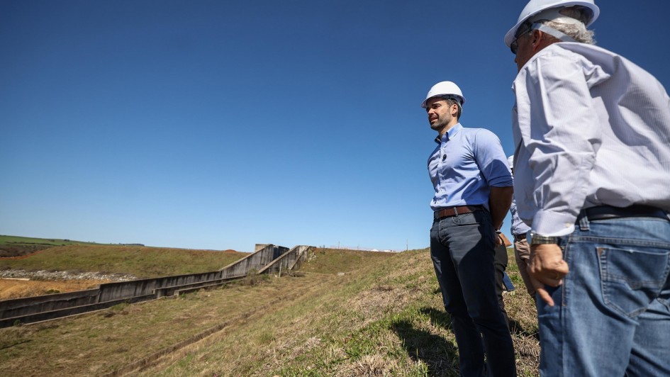 Dois homens usam capacete de proteção branco e roupas sociais casuais, observando a estrutura de uma barragem em área rural. Um deles está de camisa azul clara e calça cinza, enquanto o outro veste camisa branca e calça jeans. O céu está limpo e azul, e a cena transmite a ideia de vistoria técnica ou acompanhamento de obra. Ao fundo, é possível ver parte da estrutura de concreto da barragem e áreas de vegetação.