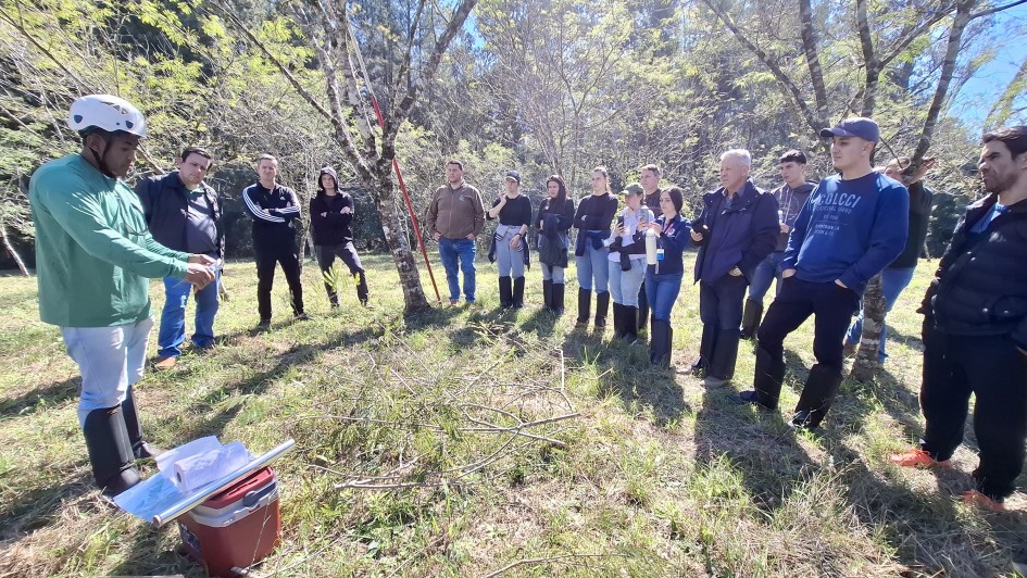 A imagem mostra um grupo de aproximadamente 17 pessoas reunidas em um ambiente ao ar livre, em uma área de vegetação com árvores e grama. O grupo está disposto em semicírculo, observando atentamente um homem à esquerda da imagem que parece estar conduzindo uma demonstração ou treinamento. Ele usa capacete branco, camisa verde, calça clara e botas de borracha. À sua frente há galhos de árvore no chão, uma caixa térmica vermelha e alguns papéis enrolados, sugerindo que a atividade está relacionada ao manejo de vegetação ou reflorestamento.

As pessoas observadoras estão vestidas de forma casual e usam botas apropriadas para trilhas ou atividades rurais. Algumas seguram garrafas ou cuias com chimarrão. O ambiente é ensolarado, com sombras projetadas pelas árvores. Ao fundo, há uma mata mais densa. A cena transmite um momento de aprendizado ou capacitação em campo, possivelmente sobre práticas ambientais ou agrícolas.