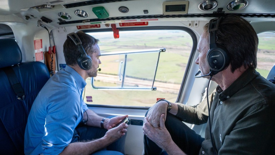 A foto, tirada de dentro de um helicóptero, mostra duas pessoas com fones de ouvido. O governador Eduardo Leite, em primeiro plano, está à direita da imagem, de perfil para a janela, com uma camisa verde-escura. O secretário de Logística e Transportes, Juvir Costella, está sentado à sua frente, com uma camisa azul-clara, segurando um objeto retangular nas mãos. Ambos olham para a paisagem exterior, que consiste em um campo verde e plano, iluminado pela luz natural. A iluminação é suave e o foco está nos passageiros e na visão lateral do helicóptero.