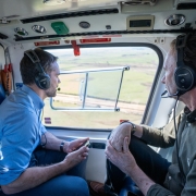A foto, tirada de dentro de um helicóptero, mostra duas pessoas com fones de ouvido. O governador Eduardo Leite, em primeiro plano, está à direita da imagem, de perfil para a janela, com uma camisa verde-escura. O secretário de Logística e Transportes, Juvir Costella, está sentado à sua frente, com uma camisa azul-clara, segurando um objeto retangular nas mãos. Ambos olham para a paisagem exterior, que consiste em um campo verde e plano, iluminado pela luz natural. A iluminação é suave e o foco está nos passageiros e na visão lateral do helicóptero.