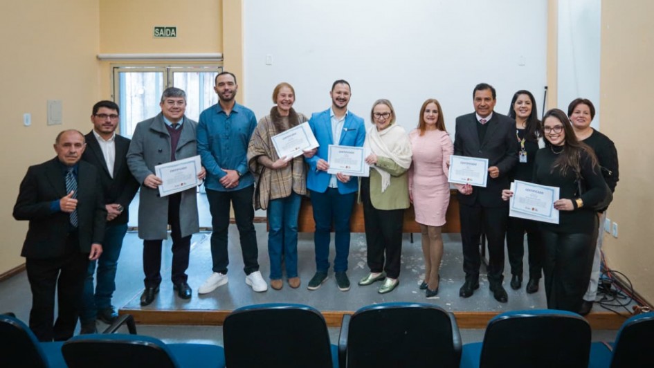 Foto posada de 12 pessoas em pé num pequeno palco num ambiente fechado. Elas usam roupas formais e sorriem. Algumas seguram os certificados, exibindo-os para a câmera.