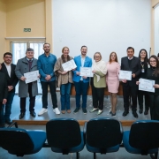 Foto posada de 12 pessoas em pé num pequeno palco num ambiente fechado. Elas usam roupas formais e sorriem. Algumas seguram os certificados, exibindo-os para a câmera.