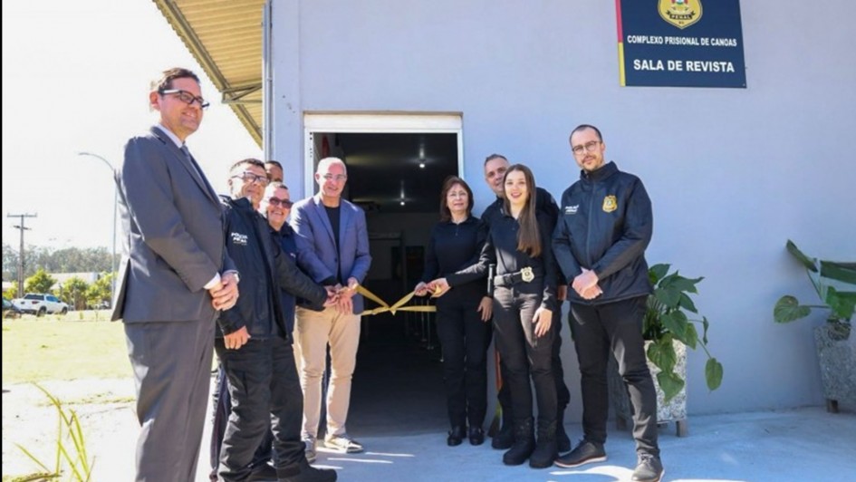 A foto mostra um grupo de nove pessoas em frente a um prédio cinza claro. A cena representa uma cerimônia de inauguração, com o grupo segurando uma faixa de fita amarela para o corte. A maioria das pessoas veste roupas pretas, que parecem ser uniformes de segurança, enquanto duas delas vestem trajes casuais de camisa e calça. O prédio possui uma placa com o texto "POLÍCIA PENAL - COMPLEXO PRISIONAL DE CANOAS - SALA DE REVISTA". Ao lado da porta, vasos com plantas verdes dão um toque de vida ao ambiente. A cena ocorre em um dia ensolarado.