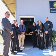 A foto mostra um grupo de nove pessoas em frente a um prédio cinza claro. A cena representa uma cerimônia de inauguração, com o grupo segurando uma faixa de fita amarela para o corte. A maioria das pessoas veste roupas pretas, que parecem ser uniformes de segurança, enquanto duas delas vestem trajes casuais de camisa e calça. O prédio possui uma placa com o texto "POLÍCIA PENAL - COMPLEXO PRISIONAL DE CANOAS - SALA DE REVISTA". Ao lado da porta, vasos com plantas verdes dão um toque de vida ao ambiente. A cena ocorre em um dia ensolarado.