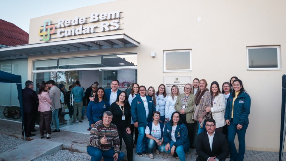 Um grupo de pessoas posa para foto em frente a um prédio de fachada clara, identificado com a placa “Rede Bem Cuidar RS”. A entrada é de vidro, e atrás dela aparecem outras pessoas no interior do espaço. Em destaque, cerca de 20 pessoas estão reunidas do lado de fora, sorrindo para a câmera. Algumas usam jalecos brancos e camisetas do programa, enquanto outras estão de casaco ou roupas sociais. No canto inferior esquerdo, um homem agachado faz sinal de positivo. O cenário é de inauguração ou evento oficial, em ambiente externo com piso de brita e calçamento.