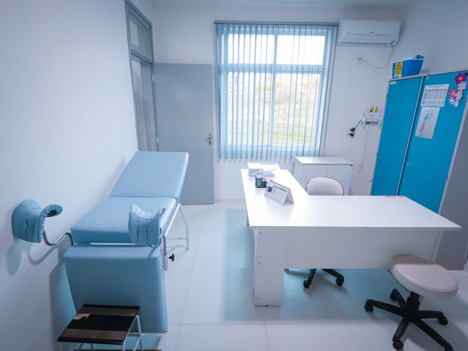 Sala de atendimento médico iluminada por luz natural que entra através de uma janela com persianas verticais. À esquerda, há uma maca ginecológica acolchoada em azul claro, com perneiras ajustáveis e um pequeno banco de apoio. No centro, uma mesa branca de escritório está organizada com papéis, pastas e suporte de crachá, acompanhada por duas cadeiras giratórias. Ao fundo, há um armário azul com enfeites coloridos e um calendário preso, além de um aparelho de ar-condicionado instalado acima da janela. O ambiente é limpo, organizado e preparado para consultas de saúde.