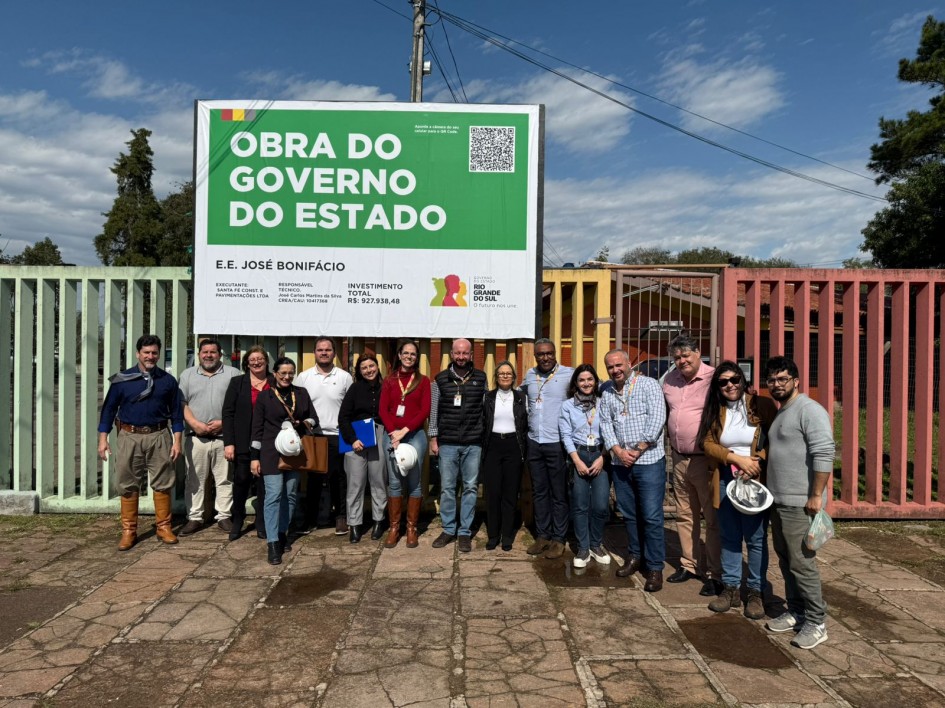 Um grupo de aproximadamente 15 pessoas está reunido em frente a um portão de escola, posando para uma foto. Atrás delas, há uma grande placa verde e branca com os dizeres “OBRA DO GOVERNO DO ESTADO” e informações sobre a execução da obra na Escola Estadual José Bonifácio, com investimento total de R$ 927.938,48. A placa também exibe o logotipo do Governo do Rio Grande do Sul. O grupo é formado por homens e mulheres, alguns segurando capacetes de segurança, todos sorrindo e olhando para a câmera. O céu está azul com algumas nuvens e o clima aparenta ser ensolarado.