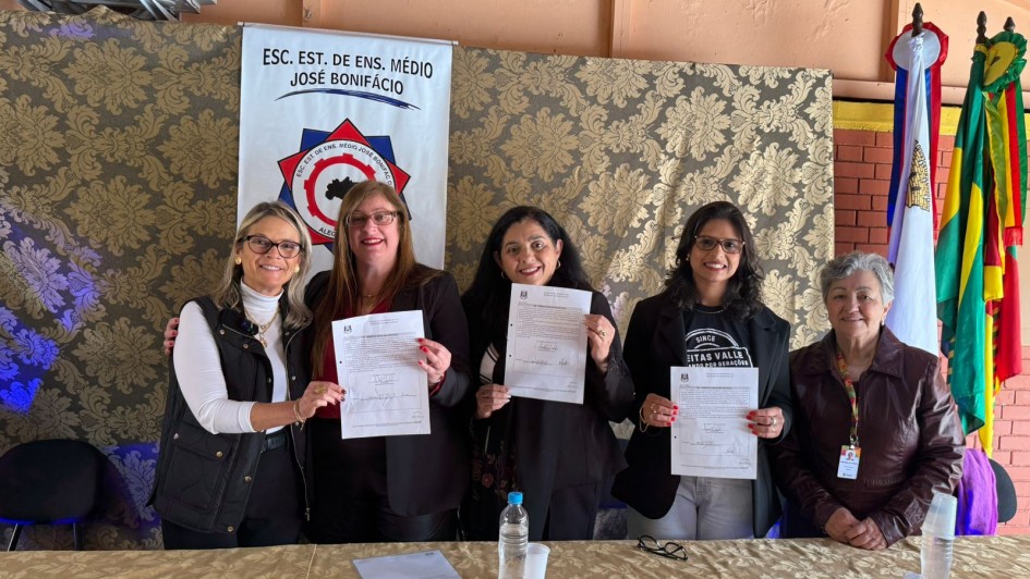 Cinco mulheres estão alinhadas atrás de uma mesa coberta por uma toalha dourada. Quatro delas, posicionadas ao centro, seguram papéis oficiais assinados e sorriem para a câmera. À esquerda, uma das mulheres veste blusa branca de gola alta e colete preto. Ao seu lado, uma mulher de óculos e cabelo ruivo veste blazer preto. No centro, outra mulher de cabelos escuros soltos veste blusa preta. À direita dela, uma jovem de óculos veste camiseta preta e blazer, e a última, à extrema direita, é uma senhora de cabelos grisalhos que veste jaqueta marrom.
Ao fundo, há um banner da Escola Estadual de Ensino Médio José Bonifácio, bandeiras do Brasil, do Rio Grande do Sul e de outros estandartes. A cena sugere um ato de assinatura ou entrega de documentos oficiais relacionados à escola.