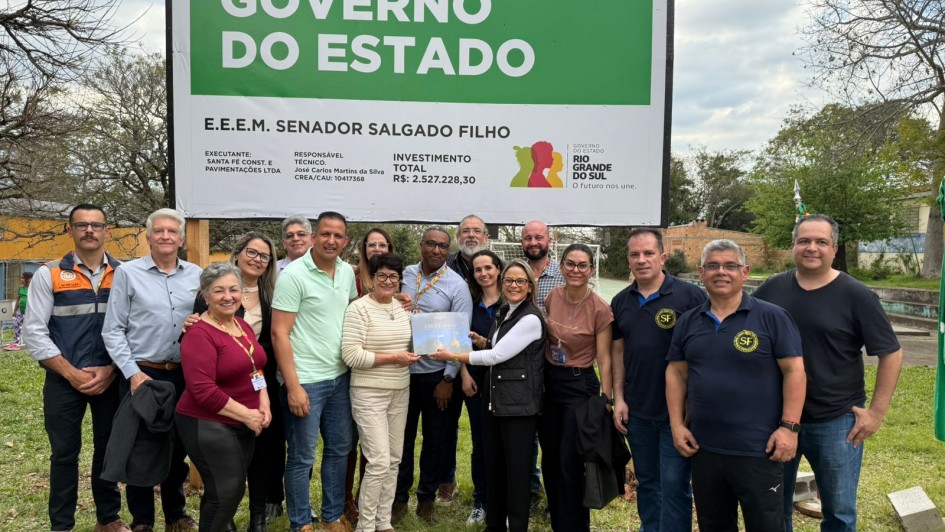 Um grupo de cerca de 18 pessoas posa sorridente em frente a uma placa verde e branca que anuncia uma obra do Governo do Estado do Rio Grande do Sul na Escola Estadual de Ensino Médio Senador Salgado Filho. A placa informa o valor do investimento, superior a R$ 2,5 milhões, e os responsáveis pela execução da obra. As pessoas estão lado a lado, algumas abraçadas, e no centro uma mulher segura um documento ou uma pasta transparente. O cenário é ao ar livre, em um gramado, com árvores sem folhas ao fundo, indicando época de outono ou inverno. Todos aparentam estar em um momento de celebração e registro oficial da obra.