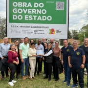 Um grupo de cerca de 18 pessoas posa sorridente em frente a uma placa verde e branca que anuncia uma obra do Governo do Estado do Rio Grande do Sul na Escola Estadual de Ensino Médio Senador Salgado Filho. A placa informa o valor do investimento, superior a R$ 2,5 milhões, e os responsáveis pela execução da obra. As pessoas estão lado a lado, algumas abraçadas, e no centro uma mulher segura um documento ou uma pasta transparente. O cenário é ao ar livre, em um gramado, com árvores sem folhas ao fundo, indicando época de outono ou inverno. Todos aparentam estar em um momento de celebração e registro oficial da obra.