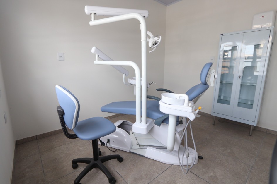 Foto de um consultório de paredes claras, com uma cadeira de dentista ao centro e um armário no fundo.