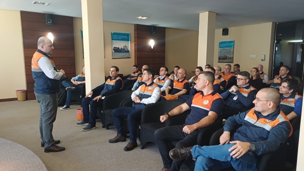 Em uma sala de auditório, um homem em pé, vestindo colete da Defesa Civil, fala para um grupo de pessoas sentadas em poltronas pretas. Os participantes, também uniformizados com camisetas azul-marinho e laranja da Defesa Civil, acompanham a apresentação com atenção. O ambiente é iluminado, com paredes em tons claros e painéis de madeira. Nas paredes laterais, há cartazes afixados.