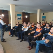 Em uma sala de auditório, um homem em pé, vestindo colete da Defesa Civil, fala para um grupo de pessoas sentadas em poltronas pretas. Os participantes, também uniformizados com camisetas azul-marinho e laranja da Defesa Civil, acompanham a apresentação com atenção. O ambiente é iluminado, com paredes em tons claros e painéis de madeira. Nas paredes laterais, há cartazes afixados.