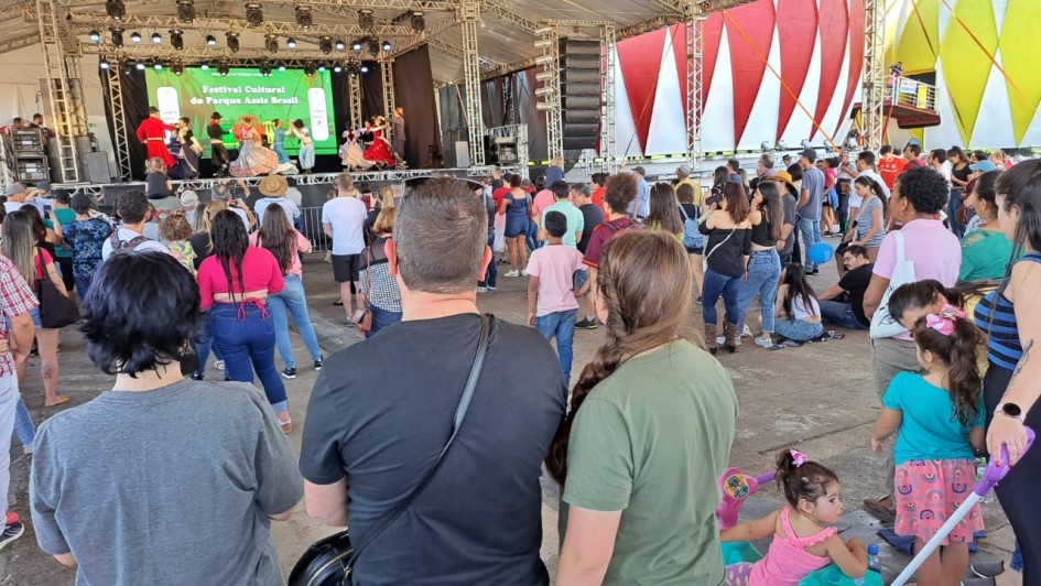Foto mostra o público de costa, várias pessoas olhando para um palco ao fundo, onde acontece uma apresentação de dança tradicionalista gaúcha. 