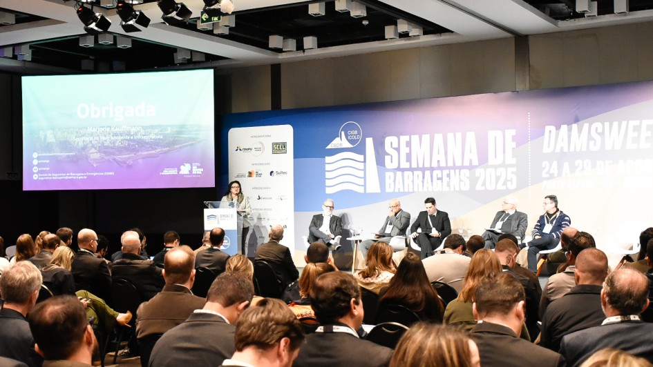 Auditório cheio, com público sentado voltado para o palco, onde ocorre um painel durante a Semana de Barragens 2025. À esquerda, uma mulher está em pé, atrás de um púlpito, realizando apresentação. Ao fundo, um telão projeta a palavra “Obrigada” e o nome de Marjorie Kauffmann, secretária estadual do Meio Ambiente e Infraestrutura. No centro do palco, cinco homens sentados participam do debate. O cenário exibe o painel azul e branco com os dizeres “Semana de Barragens 2025 – Damsweek” e a data do evento, 24 a 29 de agosto, em Porto Alegre.