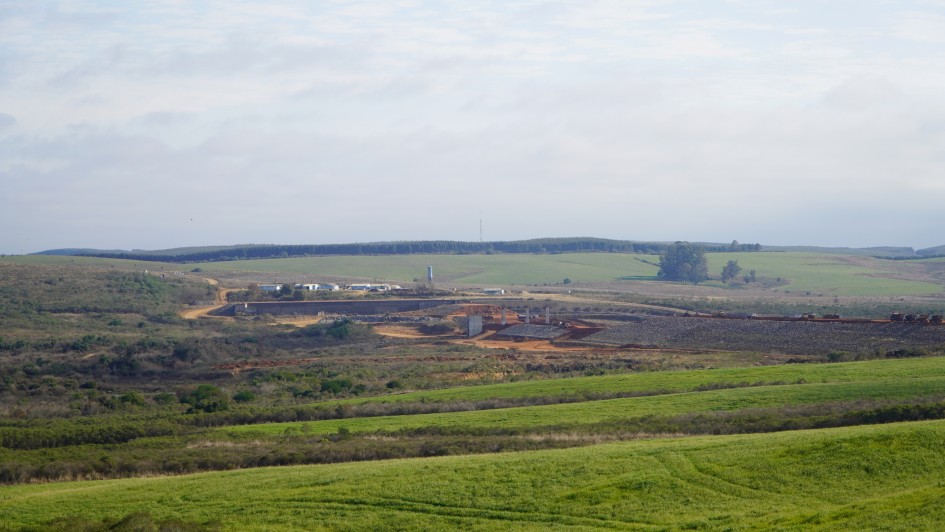 Foto externa mostra uma barragem em construção. Há grandes extensões de áreas verdes e campos próximo à obra. 