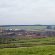 Foto externa mostra uma barragem em construção. Há grandes extensões de áreas verdes e campos próximo à obra. 