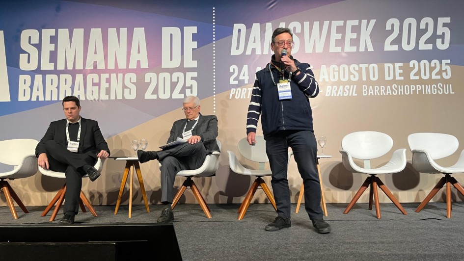 Um homem está em pé no palco, segurando um microfone e falando ao público durante a Semana de Barragens 2025 (DamsWeek 2025), realizada em Porto Alegre. Ao fundo, um grande painel exibe o nome e a data do evento: “24 a 29 de agosto de 2025”. Atrás dele, duas pessoas estão sentadas em poltronas brancas, enquanto outras cadeiras permanecem vazias. Pequenas mesas de apoio com copos de água estão dispostas ao lado das cadeiras.