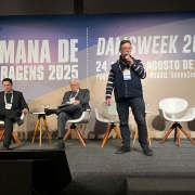 Um homem está em pé no palco, segurando um microfone e falando ao público durante a Semana de Barragens 2025 (DamsWeek 2025), realizada em Porto Alegre. Ao fundo, um grande painel exibe o nome e a data do evento: “24 a 29 de agosto de 2025”. Atrás dele, duas pessoas estão sentadas em poltronas brancas, enquanto outras cadeiras permanecem vazias. Pequenas mesas de apoio com copos de água estão dispostas ao lado das cadeiras.