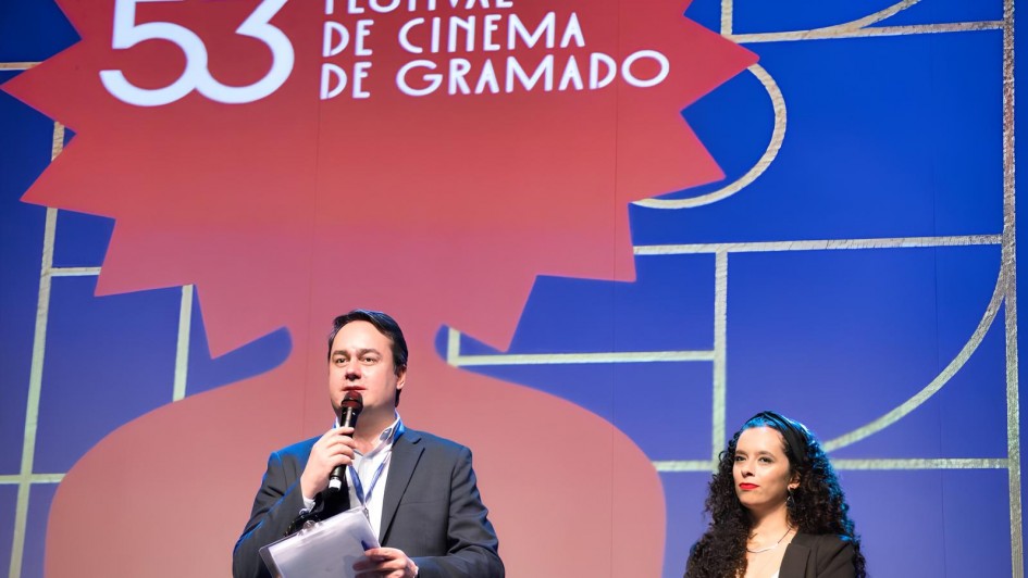 Fotografia tirada durante o 53º Festival de Cinema de Gramado. A imagem mostra um homem e uma mulher no palco do evento. O homem está à esquerda, segurando um microfone com uma das mãos e alguns papéis com a outra. Ele veste um terno escuro com camisa clara e fala ao microfone. A mulher está à direita, com cabelos cacheados soltos, longos e escuros. Ela veste um blazer preto sobre uma blusa clara e está olhando em direção ao público, com expressão séria.

Ao fundo, há um grande painel colorido em tons de azul e vermelho, com o logo do festival no topo: "53 Festival de Cinema de Gramado". O design gráfico ao fundo remete ao tradicional troféu Kikito, símbolo do evento. A iluminação é intensa, destacando os apresentadores e o logotipo do festival.