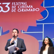 Fotografia tirada durante o 53º Festival de Cinema de Gramado. A imagem mostra um homem e uma mulher no palco do evento. O homem está à esquerda, segurando um microfone com uma das mãos e alguns papéis com a outra. Ele veste um terno escuro com camisa clara e fala ao microfone. A mulher está à direita, com cabelos cacheados soltos, longos e escuros. Ela veste um blazer preto sobre uma blusa clara e está olhando em direção ao público, com expressão séria.

Ao fundo, há um grande painel colorido em tons de azul e vermelho, com o logo do festival no topo: "53 Festival de Cinema de Gramado". O design gráfico ao fundo remete ao tradicional troféu Kikito, símbolo do evento. A iluminação é intensa, destacando os apresentadores e o logotipo do festival.