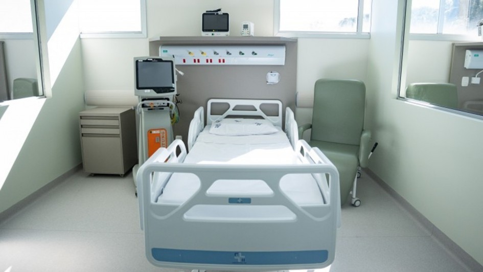 A foto mostra um quarto de hospital moderno e bem iluminado. No centro da imagem, há uma cama hospitalar branca, vazia e sem paciente, com lençóis claros. Ao lado da cama, à esquerda, está uma máquina médica cinza e branca com uma tela e tubos. Na parede acima da cama, há uma estrutura cinza com luzes de LED, tomadas, e diferentes conexões. À direita da cama, uma poltrona cinza-clara estofada. A sala é limpa, com piso branco e paredes em um tom de verde claro. Três janelas retangulares, com persianas brancas, permitem a entrada de luz natural. O ambiente transmite uma sensação de limpeza e organização.