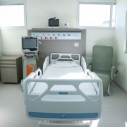 A foto mostra um quarto de hospital moderno e bem iluminado. No centro da imagem, há uma cama hospitalar branca, vazia e sem paciente, com lençóis claros. Ao lado da cama, à esquerda, está uma máquina médica cinza e branca com uma tela e tubos. Na parede acima da cama, há uma estrutura cinza com luzes de LED, tomadas, e diferentes conexões. À direita da cama, uma poltrona cinza-clara estofada. A sala é limpa, com piso branco e paredes em um tom de verde claro. Três janelas retangulares, com persianas brancas, permitem a entrada de luz natural. O ambiente transmite uma sensação de limpeza e organização.