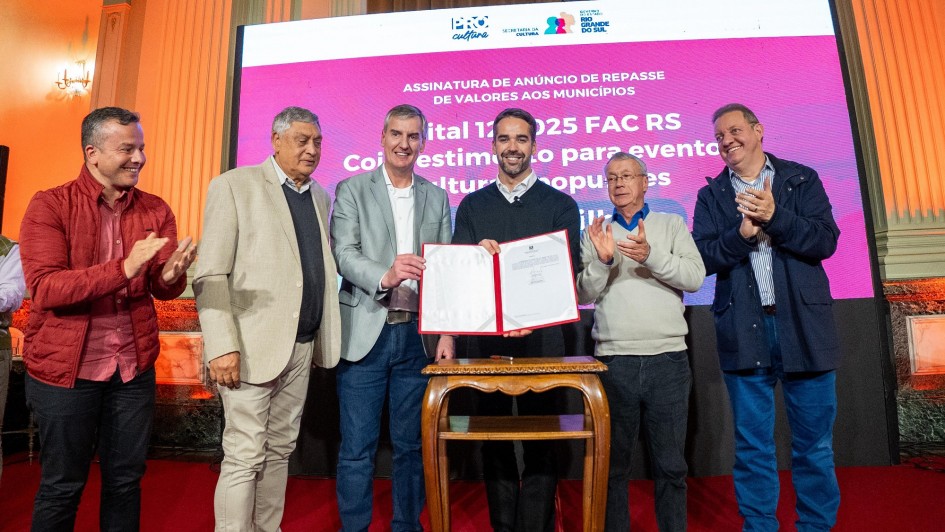 Seis homens estão em pé sobre um palco, em frente a uma grande tela eletrônica de fundo rosa com texto em branco sobre o anúncio de repasses do Fundo de Apoio à Cultura (FAC RS). No centro da imagem, dois deles seguram um documento aberto, enquanto os demais aplaudem e sorriem. O ambiente é um salão com colunas e iluminação quente, que reforça o caráter solene da cerimônia.