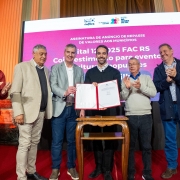 Seis homens estão em pé sobre um palco, em frente a uma grande tela eletrônica de fundo rosa com texto em branco sobre o anúncio de repasses do Fundo de Apoio à Cultura (FAC RS). No centro da imagem, dois deles seguram um documento aberto, enquanto os demais aplaudem e sorriem. O ambiente é um salão com colunas e iluminação quente, que reforça o caráter solene da cerimônia.