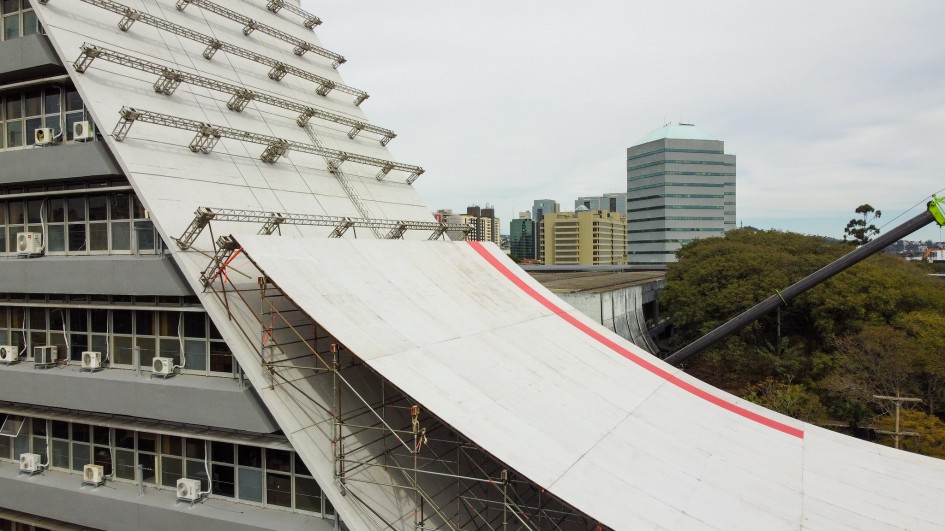 Foto da rampa, de cor branca, em processo de instalação no Caff, que aparece à esquerda do quadro. Aparece estruturas de apoio para a rampa, horizontais, sobre a superfície do prédio.