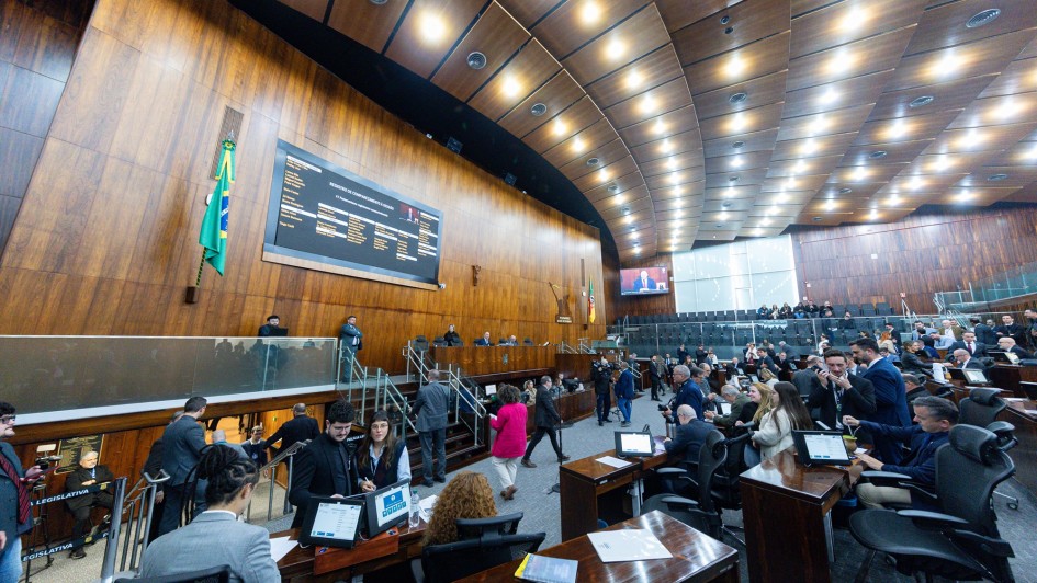 A foto mostra uma vista ampla do Plenário da Assembleia Legislativa do Rio Grande do Sul. O ambiente é dominado por painéis de madeira polida nas paredes e no teto, que é curvo e possui iluminação embutida. No centro da parede de fundo, um grande painel eletrônico exibe textos e gráficos, e abaixo dele, as bandeiras do Brasil e do Rio Grande do Sul. O plenário está cheio de pessoas, a maioria sentada em bancadas de madeira com computadores, enquanto outras circulam. A imagem também captura a presença de público na área superior, indicando um evento de grande participação.