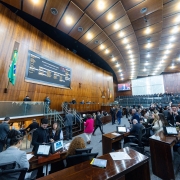 A foto mostra uma vista ampla do Plenário da Assembleia Legislativa do Rio Grande do Sul. O ambiente é dominado por painéis de madeira polida nas paredes e no teto, que é curvo e possui iluminação embutida. No centro da parede de fundo, um grande painel eletrônico exibe textos e gráficos, e abaixo dele, as bandeiras do Brasil e do Rio Grande do Sul. O plenário está cheio de pessoas, a maioria sentada em bancadas de madeira com computadores, enquanto outras circulam. A imagem também captura a presença de público na área superior, indicando um evento de grande participação.
