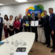 Foto em ambiente fechado de dez pessoas posando de frente, em pé. As que estão ao meio seguram o documento assinado, exibindo-o. Na parte inferior da foto, está uma mesa branca com copos.