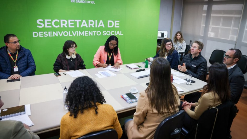 Reunião realizada em uma sala da Secretaria de Desenvolvimento Social do Rio Grande do Sul. Dez pessoas estão sentadas em torno de uma mesa retangular, participando da discussão. Ao fundo, uma parede verde com o brasão do Estado e os dizeres “Governo do Estado do Rio Grande do Sul – Secretaria de Desenvolvimento Social”. Algumas pessoas fazem anotações, outras escutam atentamente, e uma delas fala enquanto gesticula com as mãos. Sobre a mesa, há cadernos, papéis, copos e xícaras.