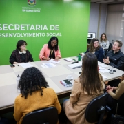 Reunião realizada em uma sala da Secretaria de Desenvolvimento Social do Rio Grande do Sul. Dez pessoas estão sentadas em torno de uma mesa retangular, participando da discussão. Ao fundo, uma parede verde com o brasão do Estado e os dizeres “Governo do Estado do Rio Grande do Sul – Secretaria de Desenvolvimento Social”. Algumas pessoas fazem anotações, outras escutam atentamente, e uma delas fala enquanto gesticula com as mãos. Sobre a mesa, há cadernos, papéis, copos e xícaras.