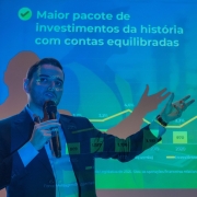 A foto mostra o vice-governador Gabriel Souza, de terno e camisa azuis, falando para uma plateia. Ele está com um microfone na mão esquerda, e sua mão direita está levantada, com os dedos abertos, enquanto ele se expressa. Atrás dele, um telão iluminado exibe um gráfico de barras verdes e uma linha amarela em ascensão. O texto principal no telão é "Maior pacote de investimentos da história com contas equilibradas". Acima do gráfico de barras, há outro texto: "Governo do RS alcança maior índice de investimentos em 25 anos". As informações indicam o crescimento da economia.