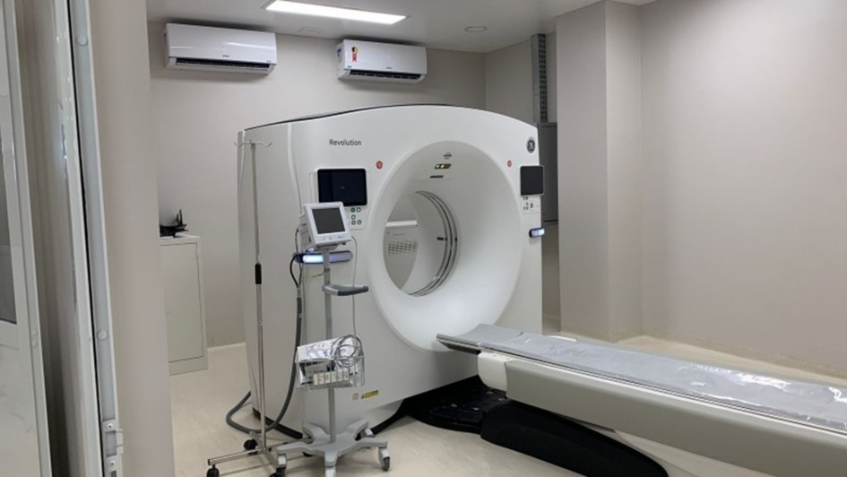 A foto mostra um aparelho médico de grande porte em uma sala de hospital, provavelmente de ressonância magnética. O aparelho principal é branco, tem um formato circular. Uma maca deslizante se projeta da parte circular, com um colchonete cinza sobre ela. Há outros equipamentos médicos menores ao lado, como um monitor em um carrinho. As paredes são de cor creme clara, o chão é cinza.