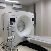 A foto mostra um aparelho médico de grande porte em uma sala de hospital, provavelmente de ressonância magnética. O aparelho principal é branco, tem um formato circular. Uma maca deslizante se projeta da parte circular, com um colchonete cinza sobre ela. Há outros equipamentos médicos menores ao lado, como um monitor em um carrinho. As paredes são de cor creme clara, o chão é cinza.