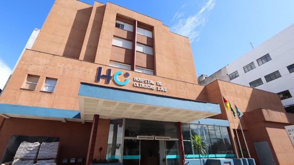 A foto mostra a fachada de um prédio de tijolos vermelhos, identificado como Hospital de Clínicas de Ijuí. O nome do hospital está em letras azuis-claras sobre a entrada principal, que possui uma cobertura azul e janelas de vidro. Acima da entrada, as letras "HC" em azul se destacam. Bandeiras, incluindo a do Brasil e do Rio Grande do Sul, estão penduradas em um mastro. O céu, com algumas nuvens, é de um azul forte. À direita, um prédio vizinho de cor branca é visível. A iluminação é natural, e a imagem é nítida.