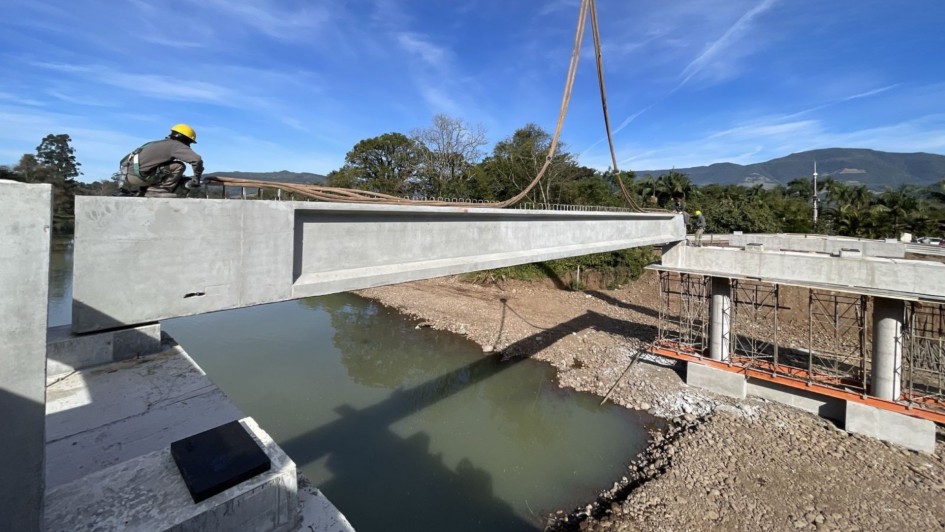 A foto mostra a construção de uma ponte sobre o Rio Três Forquilhas. Uma grua com um gancho içado está em primeiro plano, sustentando uma grande viga de concreto cinza. Um operário, usando capacete amarelo, está agachado na viga, controlando o processo. A estrutura de concreto se estende sobre o rio, cujas águas esverdeadas e turvas refletem o céu. O ambiente é iluminado pelo sol, e no fundo, é possível ver árvores verdes e montanhas sob um céu azul. A cena capta a complexidade e a escala da obra em andamento.