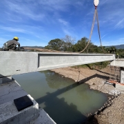 A foto mostra a construção de uma ponte sobre o Rio Três Forquilhas. Uma grua com um gancho içado está em primeiro plano, sustentando uma grande viga de concreto cinza. Um operário, usando capacete amarelo, está agachado na viga, controlando o processo. A estrutura de concreto se estende sobre o rio, cujas águas esverdeadas e turvas refletem o céu. O ambiente é iluminado pelo sol, e no fundo, é possível ver árvores verdes e montanhas sob um céu azul. A cena capta a complexidade e a escala da obra em andamento.