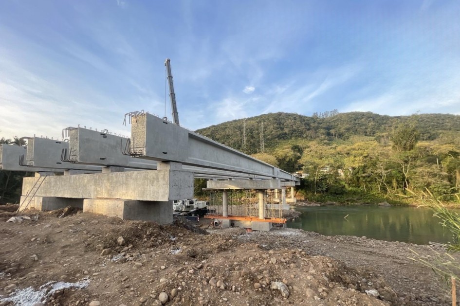 A foto mostra a nova ponte sobre o Rio Três Forquilhas em um estágio avançado de construção, vista de um ângulo baixo. Várias vigas de concreto cinza estão apoiadas em pilares robustos. Um guindaste, visível ao fundo, sugere que as obras estão em progresso. A paisagem ao redor é composta por um rio com água esverdeada, a margem de terra e rochas, e uma densa vegetação verde nas encostas. A iluminação natural do sol poente cria um brilho dourado no céu, contrastando com o cinza do concreto e o verde da natureza.