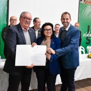 Dois homens e uma mulher estão de pé, posando para a câmera. Eles seguram um documento e sorriem. Ao fundo, há diversas pessoas e uma mesa com cadeiras. 