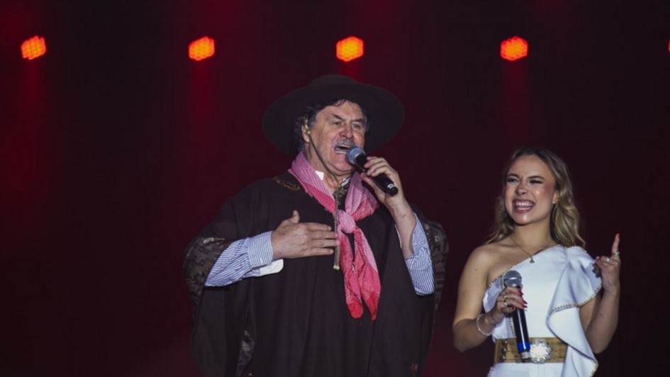 Um homem e uma mulher cantam no palco durante um show. O homem veste traje tradicional gaúcho, incluindo chapéu preto, poncho escuro, camisa listrada e lenço vermelho no pescoço. Ele segura um microfone e canta com expressão séria. Ao lado, a mulher usa vestido branco de um ombro só, com cinto dourado, e também segura um microfone, sorrindo enquanto canta. Ao fundo, luzes laranjas iluminam o cenário.
