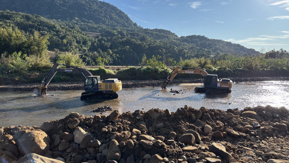 A imagem mostra dois tratores de escavação, um da marca John Deere (amarelo) e outro da marca Case (preto), operando na margem e dentro de um rio. O trator John Deere está mais perto da margem, com a caçamba abaixada na água, enquanto o trator Case está mais no meio do rio. Na frente da imagem há um monte de pedras e terra. O cenário inclui uma paisagem montanhosa com vegetação densa no fundo. O dia está ensolarado com céu azul.