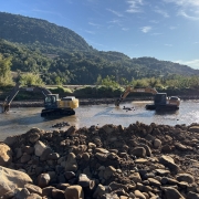 A imagem mostra dois tratores de escavação, um da marca John Deere (amarelo) e outro da marca Case (preto), operando na margem e dentro de um rio. O trator John Deere está mais perto da margem, com a caçamba abaixada na água, enquanto o trator Case está mais no meio do rio. Na frente da imagem há um monte de pedras e terra. O cenário inclui uma paisagem montanhosa com vegetação densa no fundo. O dia está ensolarado com céu azul.