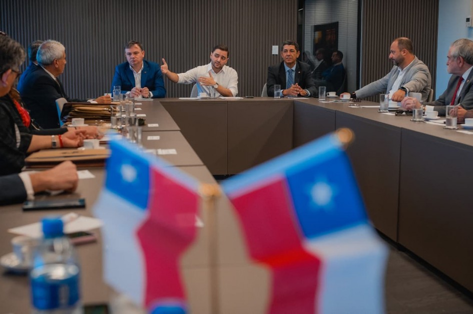 Foto de uma mesa de reuniões em formato de U, de cor marrom claro. Os participantes estão sentados junto à mesa e olham para Gabriel, que está na ponta, no centro da foto e gesticula com o braço esticado enquanto fala. No primeiríssimo plano, na metade inferior do quadro, aparecem duas pequenas bandeiras do Chile, borradas pela proximidade da câmera, composta pelas cores vermelho, azul e branco.