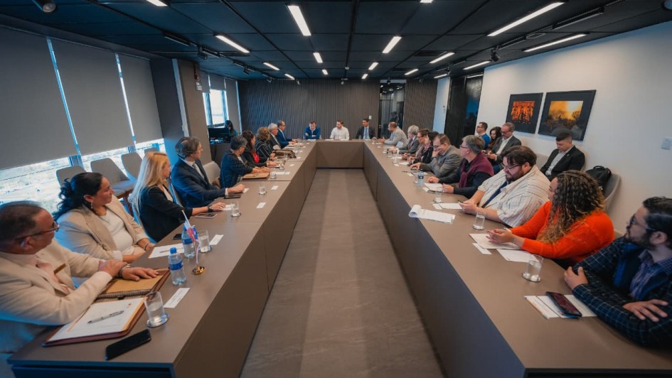 Foto geral de uma sala com uma grande mesa em formato de U, de cor marrom. Há mais de 20 pessoas sentadas junto à mesa. O ambiente é formal.
